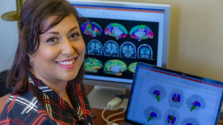 Arpana Church sitting at a desk with colorful brain imaging scans displayed on a laptop and computer screen.