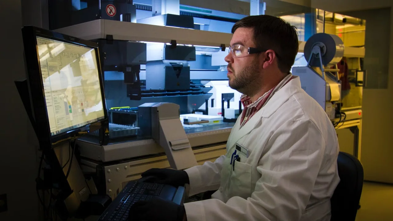 Scientist in lab coat operating laboratory automation equipment using a computer workstation.