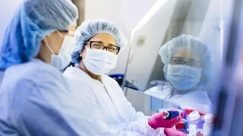Two scientists in protective gear working in a lab hood, one holding a pipette.