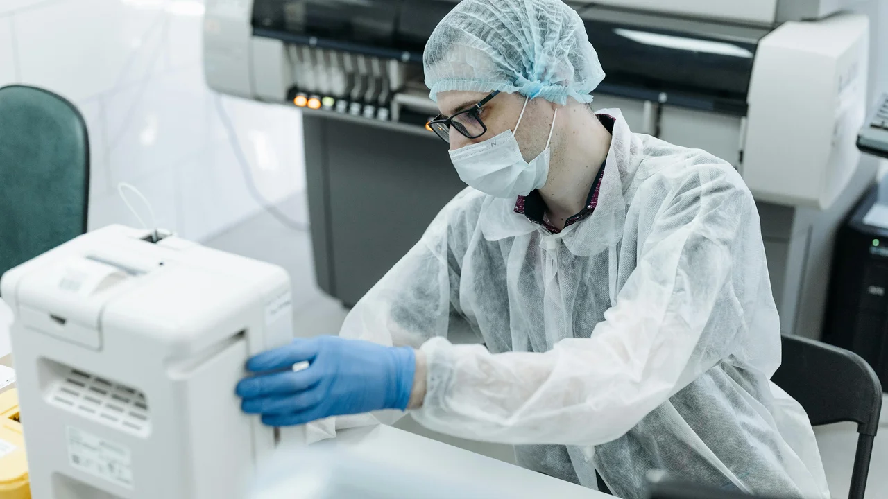 A person wearing protective clothing, a face mask, and gloves operates laboratory equipment in a clinical or research lab setting.
