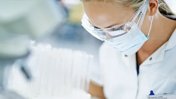 A scientist wearing a mask and goggles studies test tubes. 