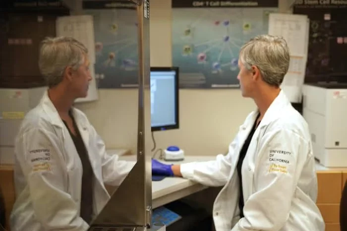 Woman in lab coat working at computer in laboratory, reflected in glass divider.