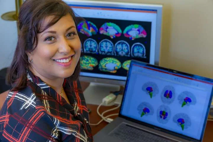 Arpana Church sitting at a desk with colorful brain imaging scans displayed on a laptop and computer screen.