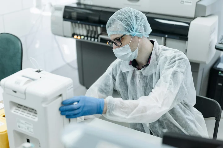 A person wearing protective clothing, a face mask, and gloves operates laboratory equipment in a clinical or research lab setting.