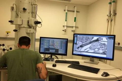 Researcher operating a transmission electron microscope while viewing high-resolution cell images on dual monitors.